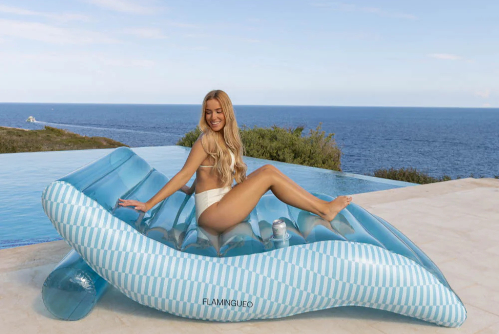 Blonde vrouw op een blauwe floatie lounge stoel lang het zwembad met de zee op de achtergrond
