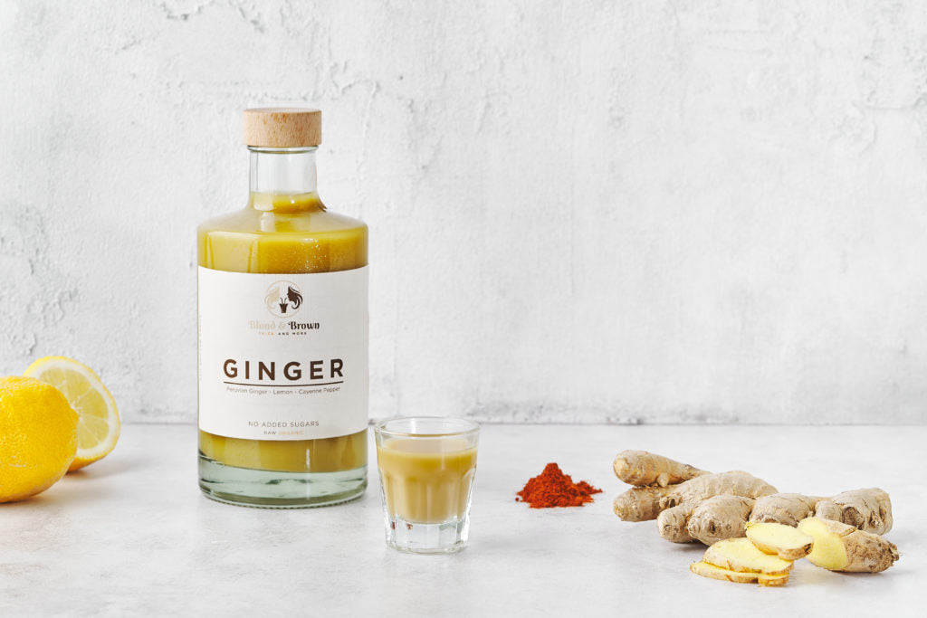 Fles gembersap van blond en brown met een shotglaasje voor, sinaasappelen links, cayenne peper en gember rechts van de fles.